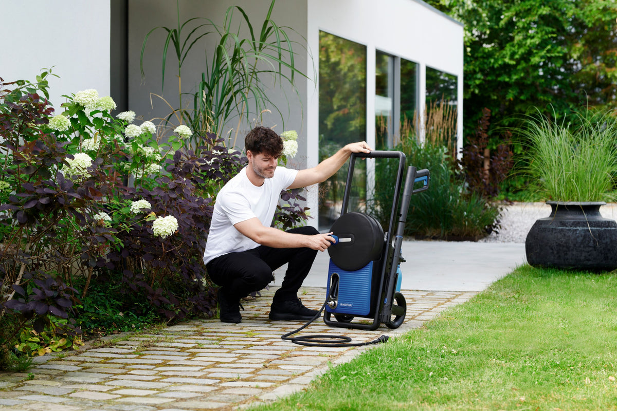 Man kneels on stone path, connecting hose to blue pressure washer near lush garden and modern home.