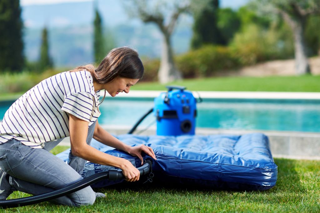 Blue wet and dry vacuum cleaner by Nilfisk-ALTO, next to a woman inflating a blue air mattress on green grass.