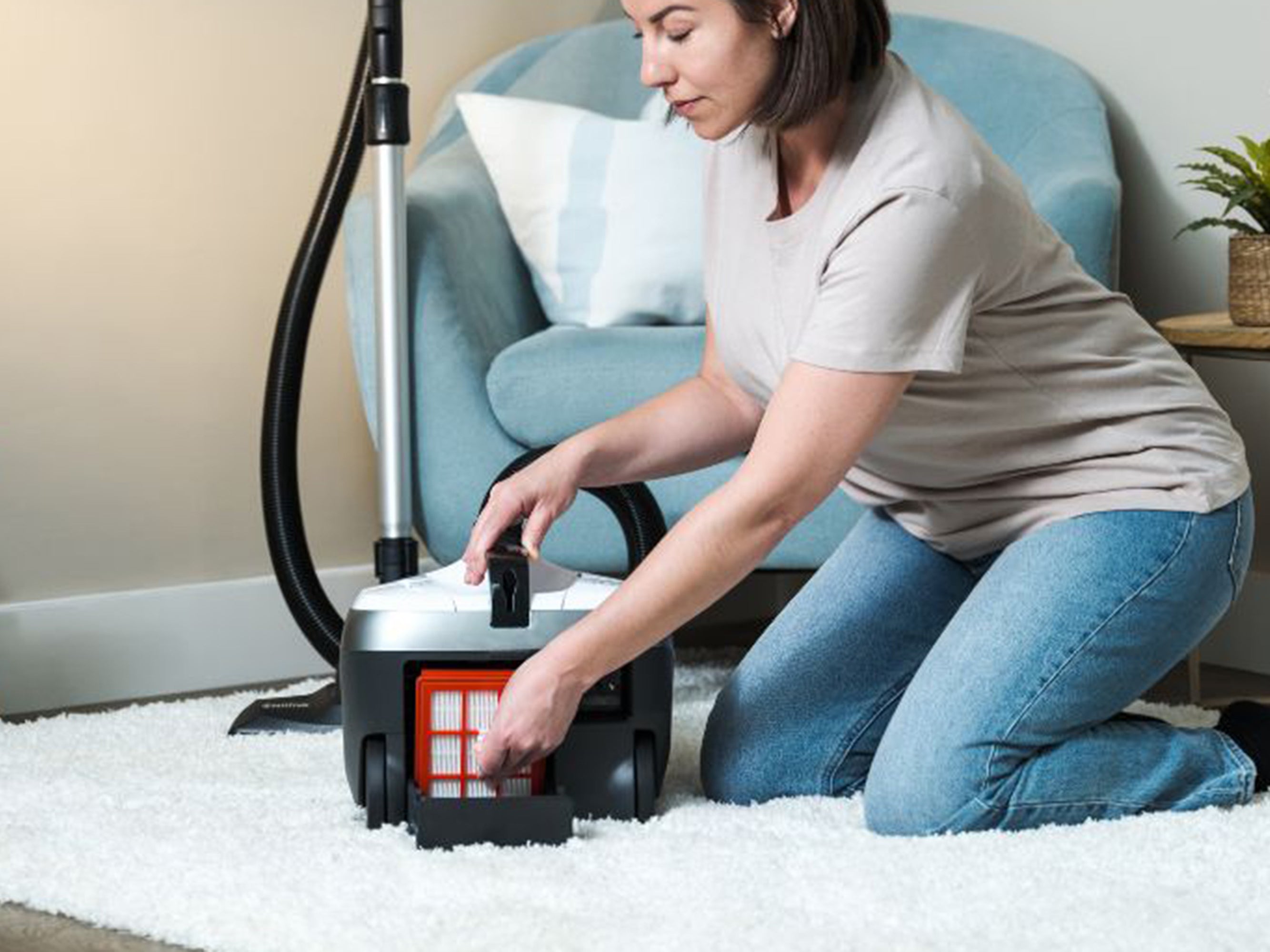 Woman kneeling on white rug, carefully removing filter from upright vacuum cleaner in cozy living room.