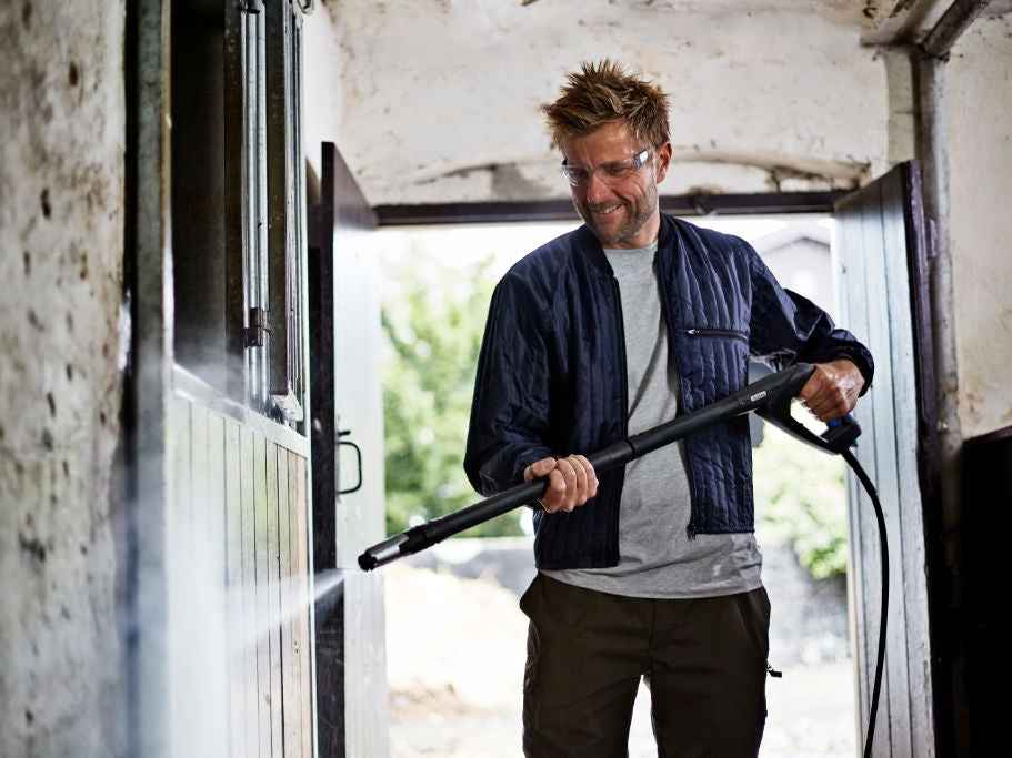 Man using high-pressure washer to clean horse stable walls, wearing safety glasses and smiling.