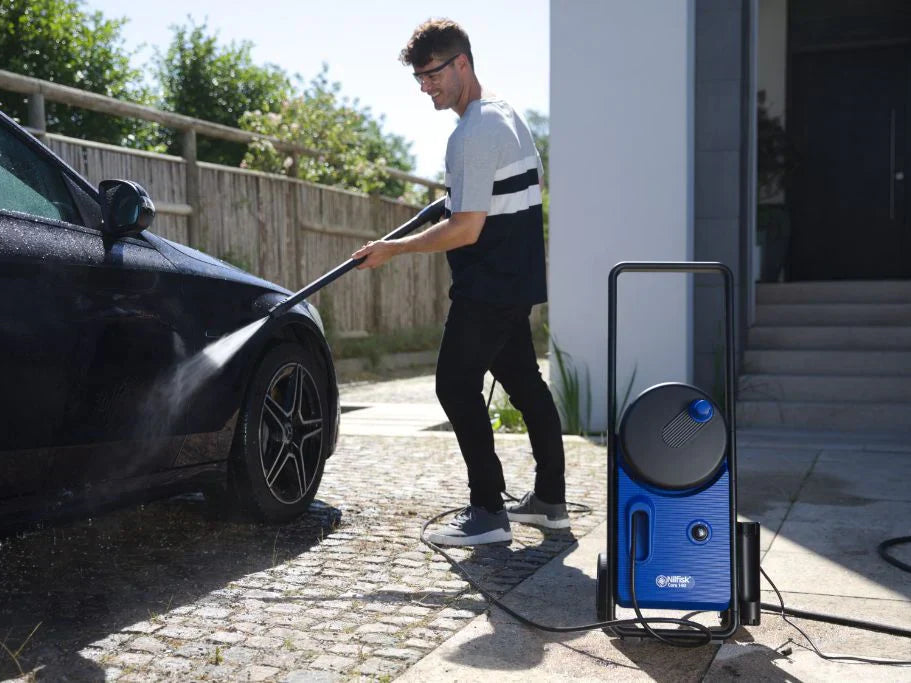 Man using blue pressure washer to clean black car in sunny driveway, water spraying from nozzle.
