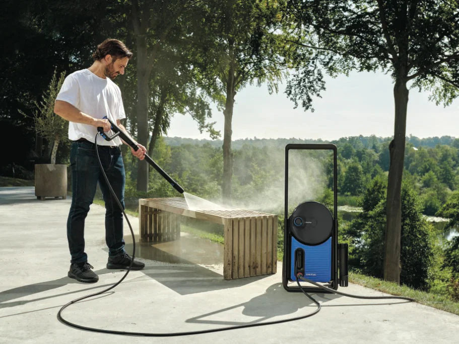 Man cleaning wooden bench with blue pressure washer in sunny garden, mist spraying, trees in background.