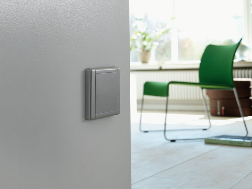 Modern silver light switch on gray wall, bright living room with green chair and sunlight streaming through window.