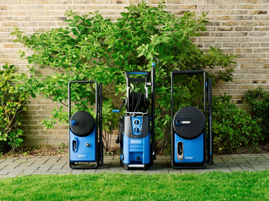 Three blue high-pressure washers lined up on a patio, ready for outdoor cleaning, set against a brick wall and green shrubs.