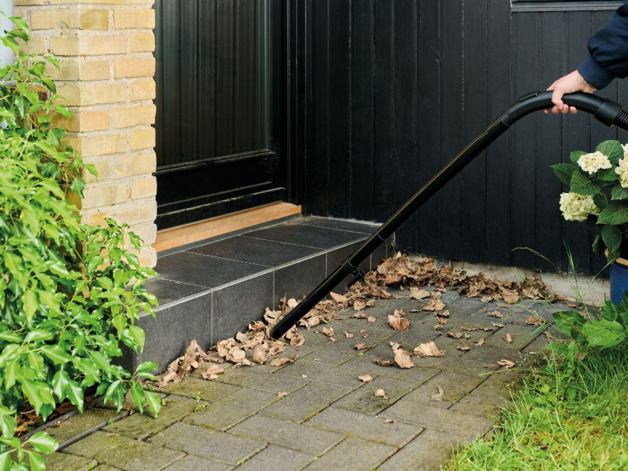 Person using wet/dry vacuum to clean fallen leaves from stone patio near house entrance, surrounded by greenery and flowers.