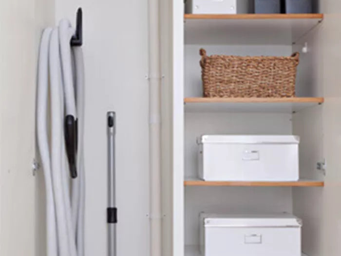 Neatly organized closet with vacuum hose hung on hook, shelves holding white boxes, folders, and a wicker basket.