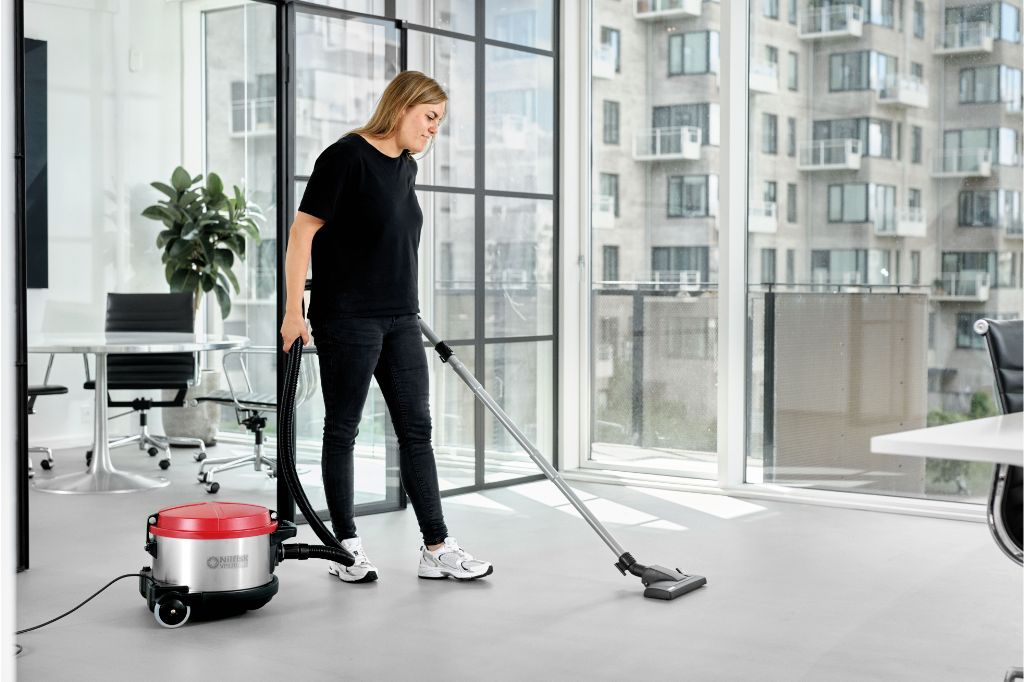 Vacuum cleaner by Nilfisk is used on a gray floor in front of a large window.