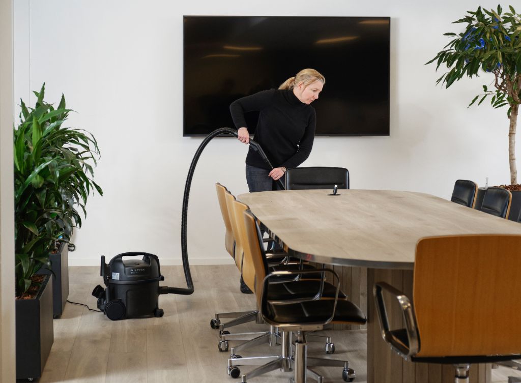 Black canister vacuum cleaner with HEPA filter and 800 W motor, by Nilfisk, in office setting with wooden table and chairs, side view