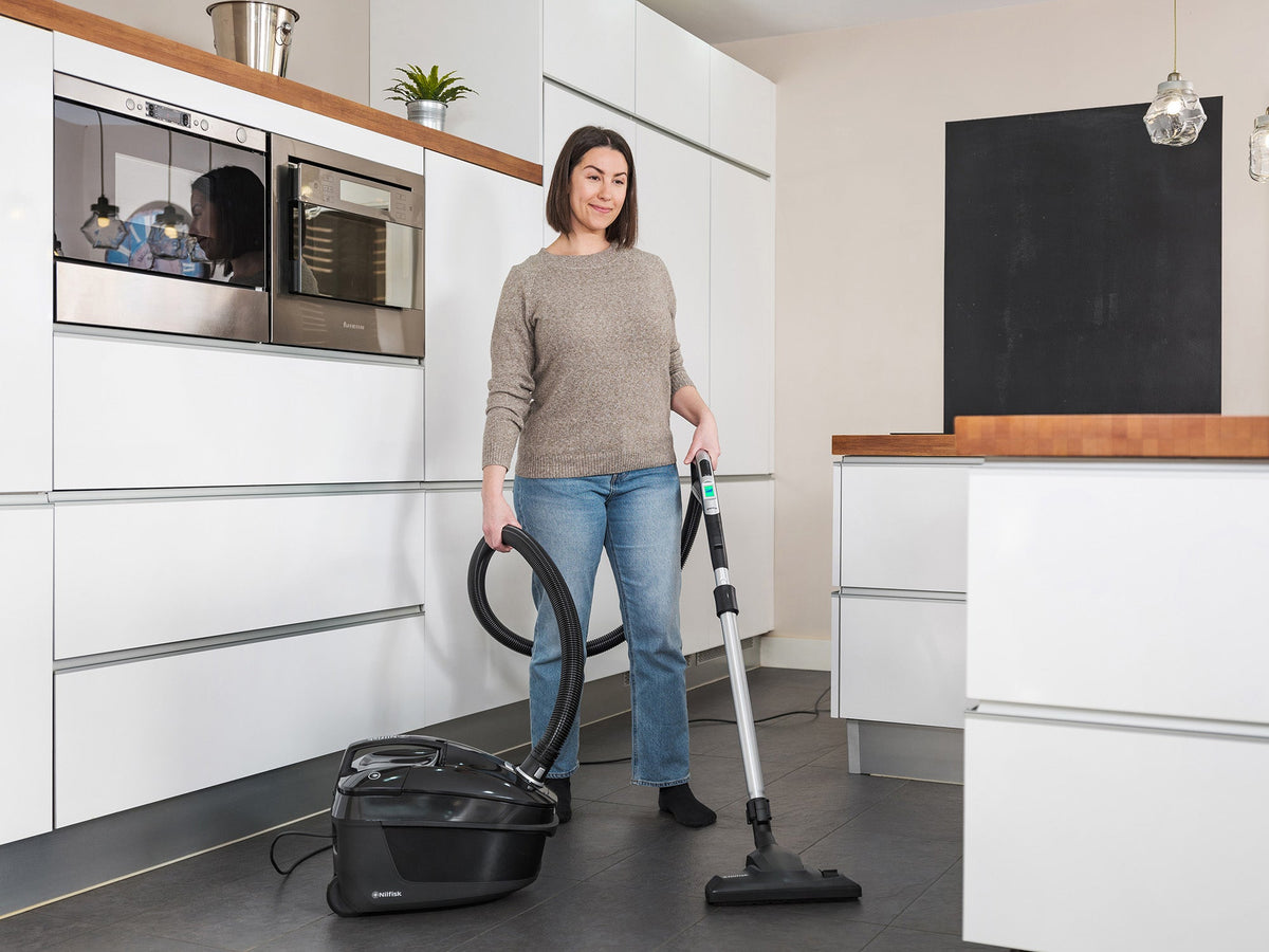 Black canister vacuum cleaner by Nilfisk in a modern kitchen, with woman wearing a sweater and jeans.

