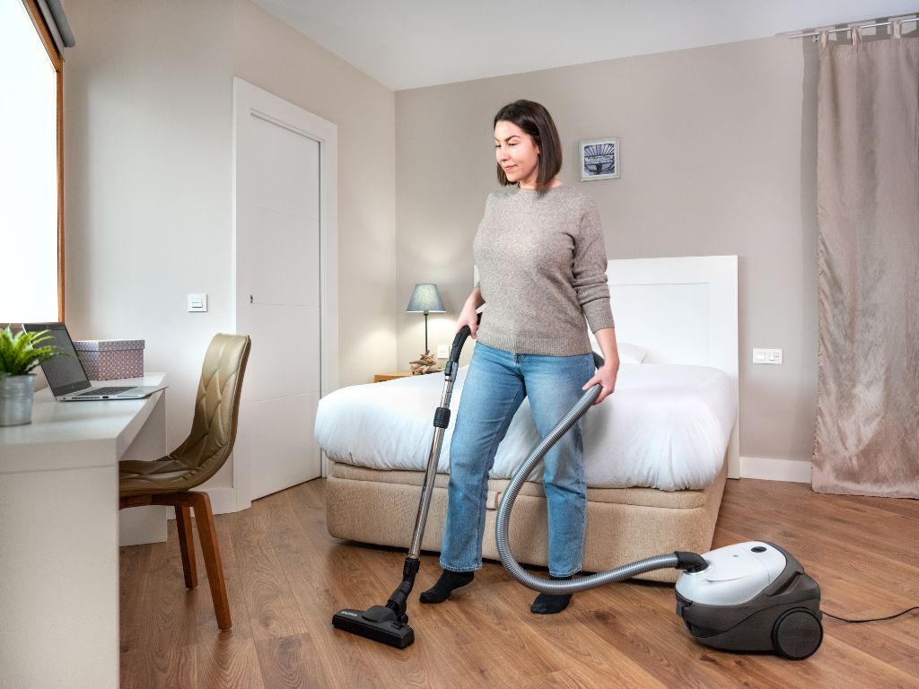 White and gray canister vacuum cleaner by ES One Allergy, used on hardwood floor in bedroom, with flexible hose and floor nozzle, lifestyle view
