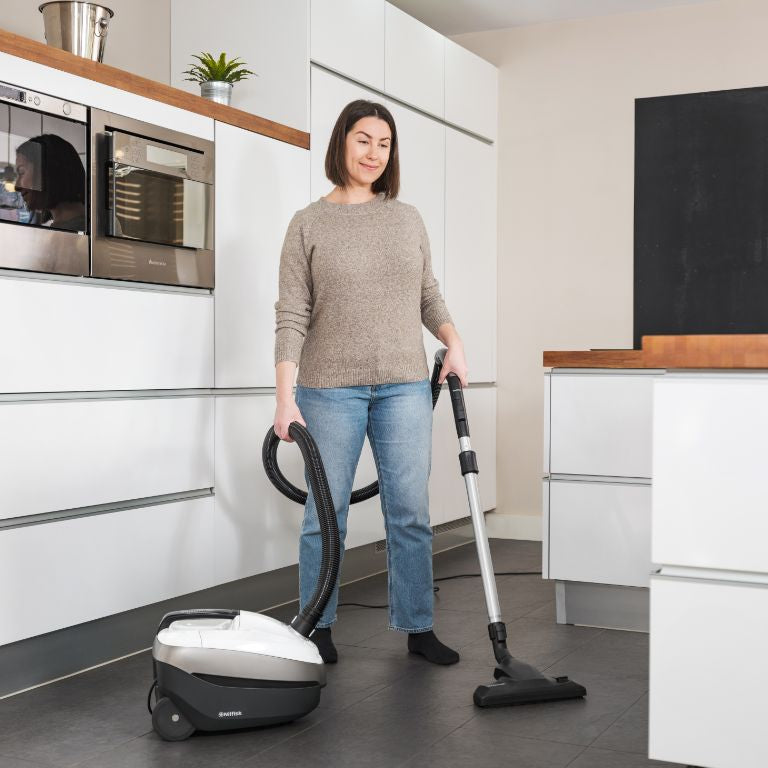Bagless vacuum cleaner by Nilfisk being used in a modern white kitchen, with dark gray floors and a woman vacuuming.