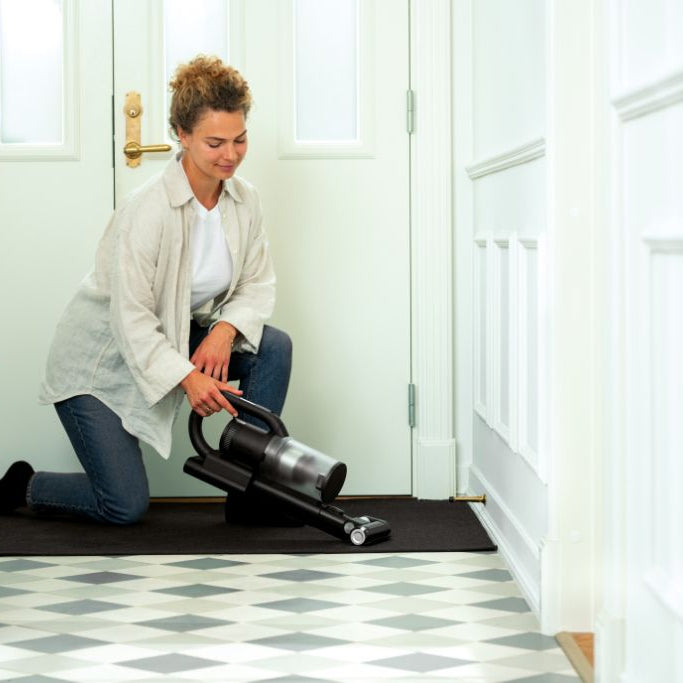 Vacuum cleaner accessory by S1 being used on a dark floor mat, in a tiled hallway with a fair-skinned woman.