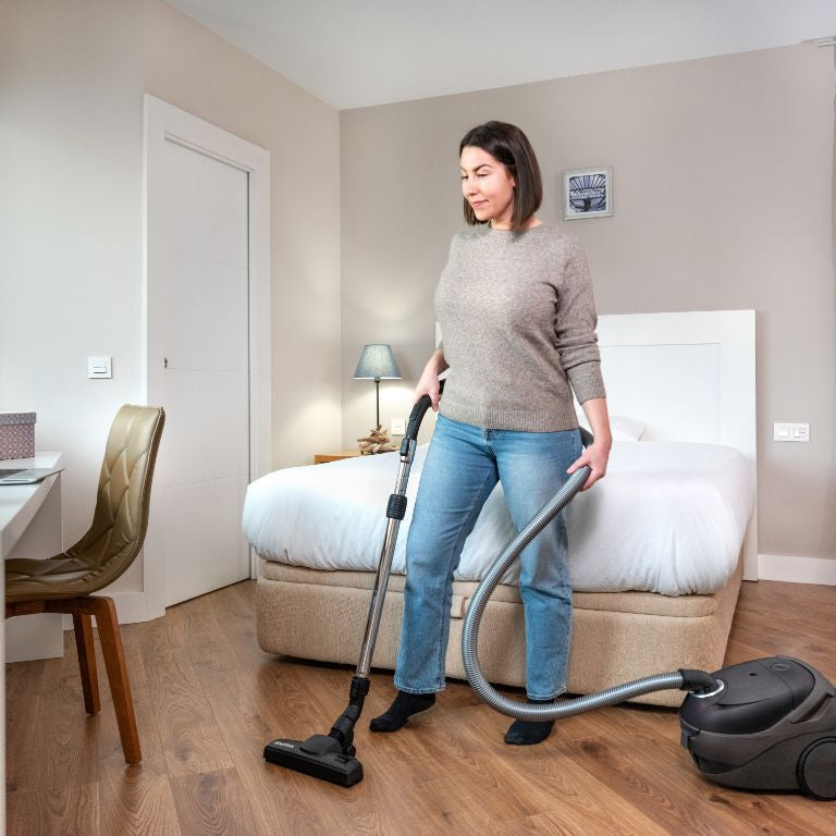Black canister vacuum cleaner by ES One Pet Care 750 W, with flexible hose and floor nozzle, used on wooden flooring in bedroom setting