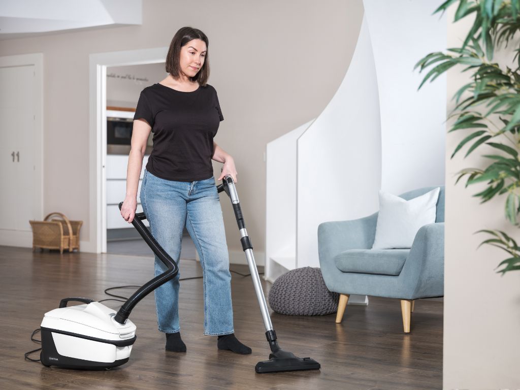 White cylinder vacuum cleaner by Nilfisk on dark wood flooring, woman vacuuming.