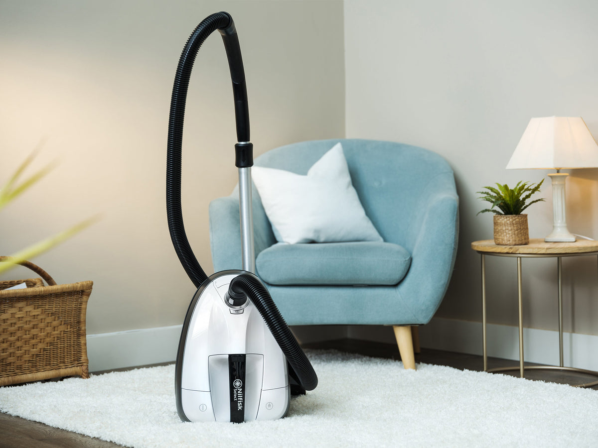 White and black vacuum cleaner by Nilfisk standing on a white fluffy rug in a staged room.
