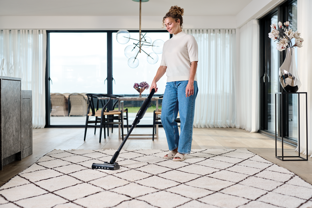 Black cordless stick vacuum cleaner with docking station by Move, shown in use on patterned rug in bright living room, side view.