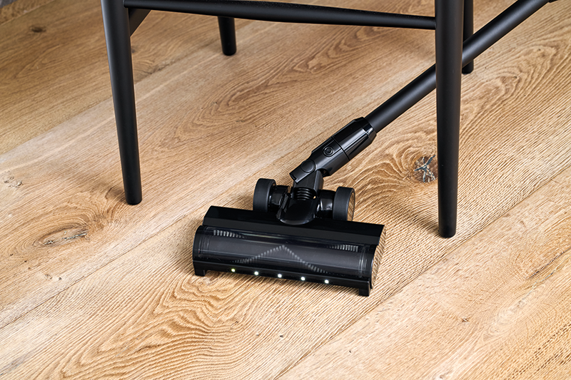 Black cordless vacuum cleaner with docking station, 150 W, on wooden floor, side view, LED lights on brush head