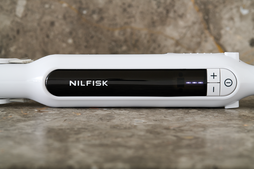 White handheld vacuum cleaner by Nilfisk with black grip panel, power buttons, and blue LED display, side view on stone surface
