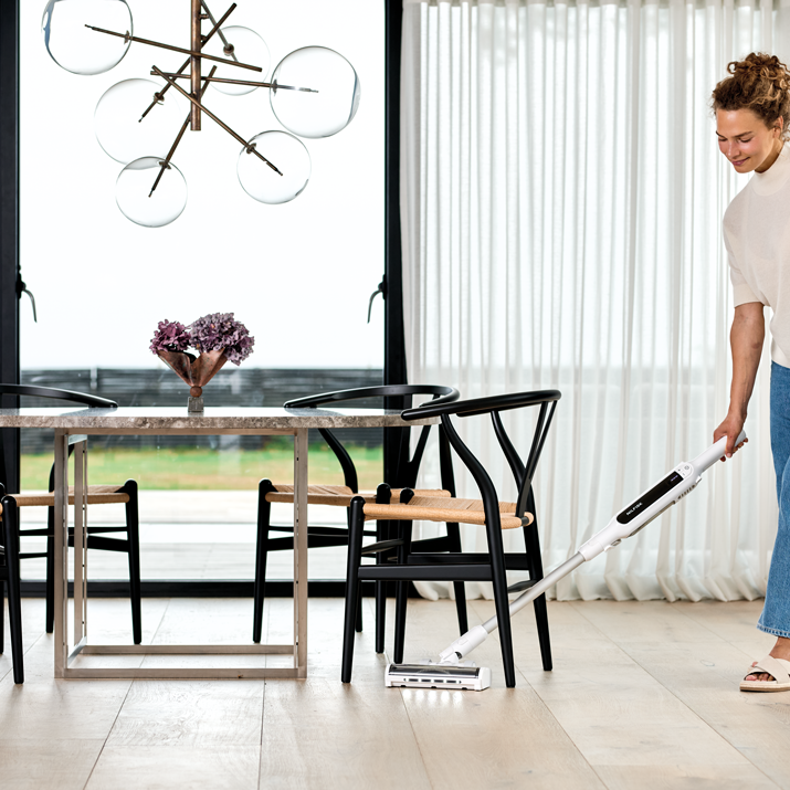 White cordless stick vacuum with docking station by Move, shown in use on light wood flooring in modern dining area, side view.