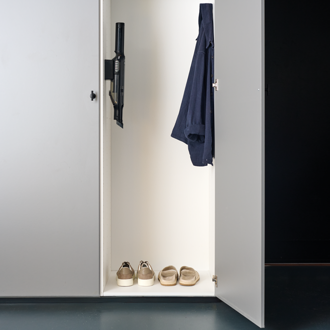Black handheld vacuum cleaner by Spot mounted on white cabinet door, sleek design, minimalist hallway setting, shoes and coat visible inside.