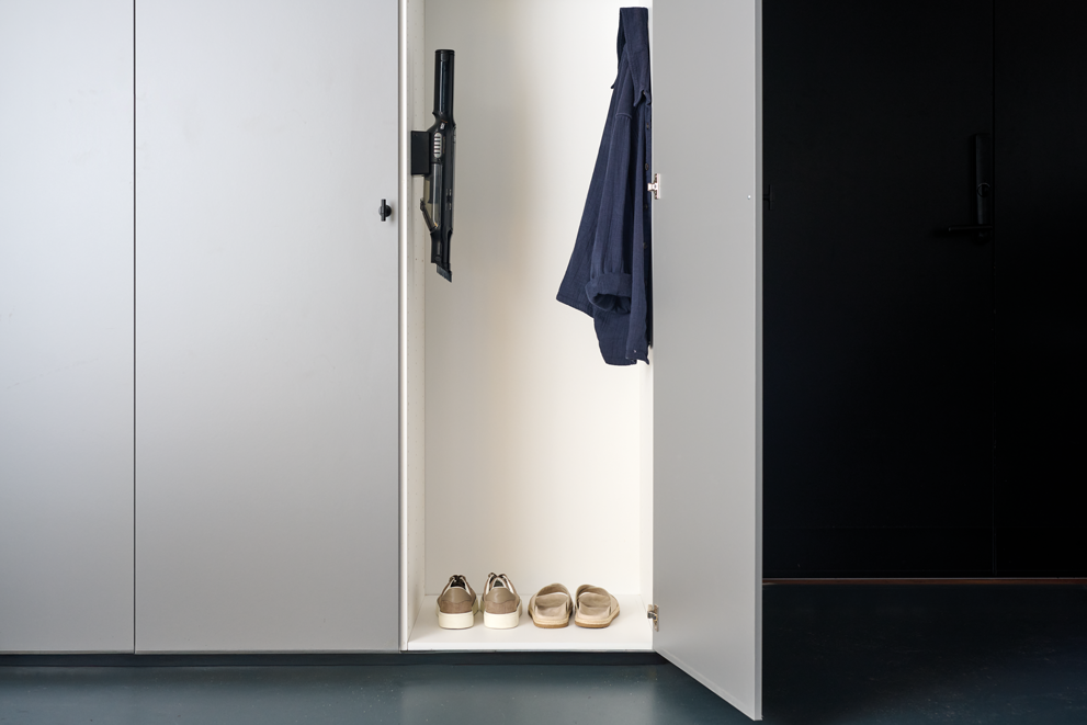 Black handheld vacuum cleaner by Spot mounted on white cabinet door, sleek design, minimalist hallway setting, shoes and coat visible inside.