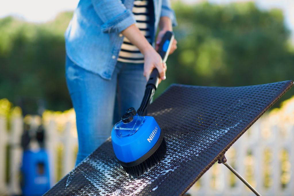 Rotary brush head accessory by Nilfisk cleaning a woven outdoor lounge chair, with denim jacket and striped shirt in the background.