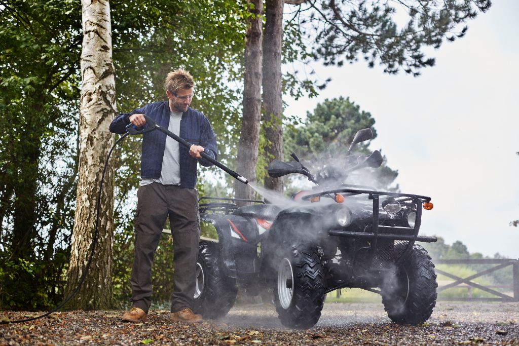 Black high-pressure washer with long hose and spray nozzle in use, cleaning ATV outdoors, side view, water mist visible