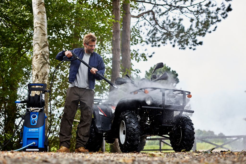 High pressure washer by Nilfisk spraying an ATV, with a man operating the wand outdoors near trees.