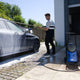 Blue and black high-pressure washer by Core 140 in outdoor use, spraying soapy water on a dark car, side view on paved driveway.