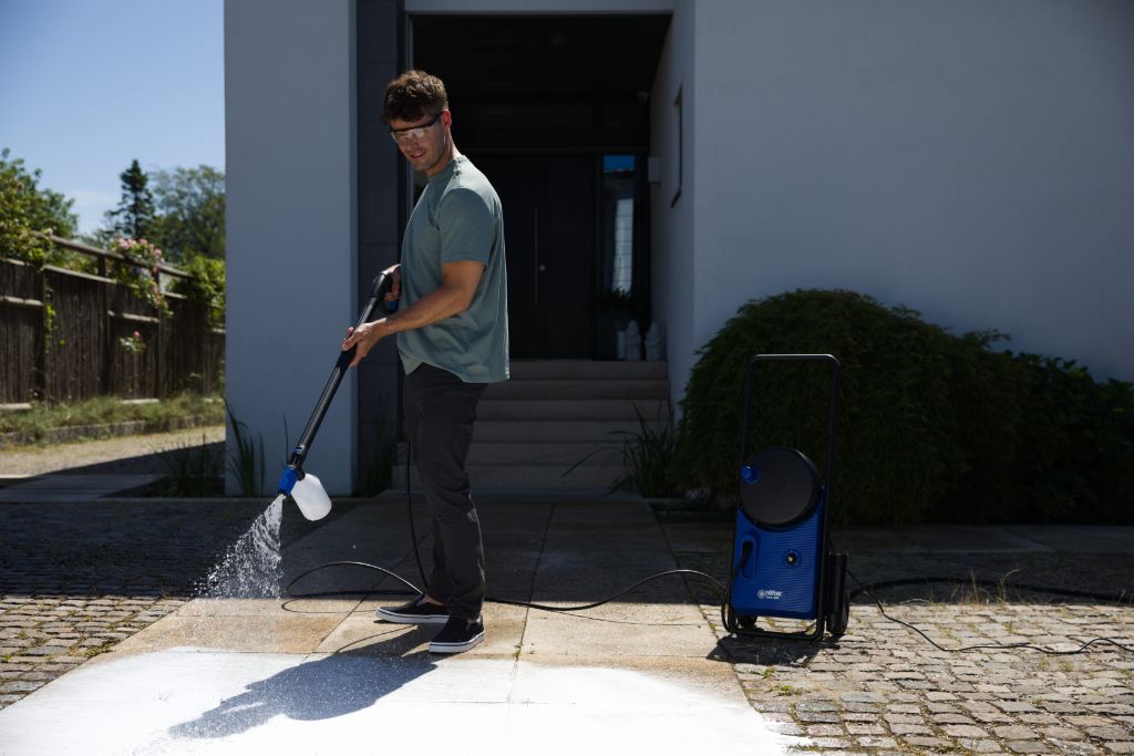 Blue and black high-pressure washer with hose and spray gun, in use on cobblestone patio, side view, lifestyle shot