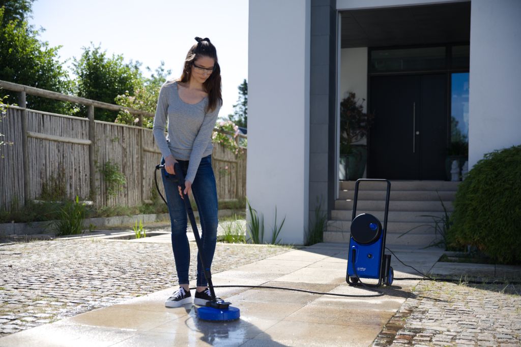 High pressure washer by Nilfisk with blue housing on a paved patio while in use by a woman.