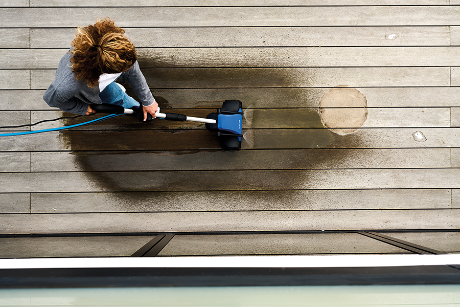 Blue and black terrace cleaner by Bosch in use on wooden deck, overhead view showing water spray and cleaning path