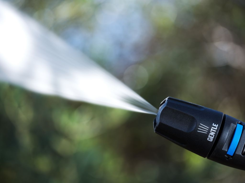 Black pressure washer nozzle in gentle mode, by Nilfisk, spraying water against a blurred nature background.