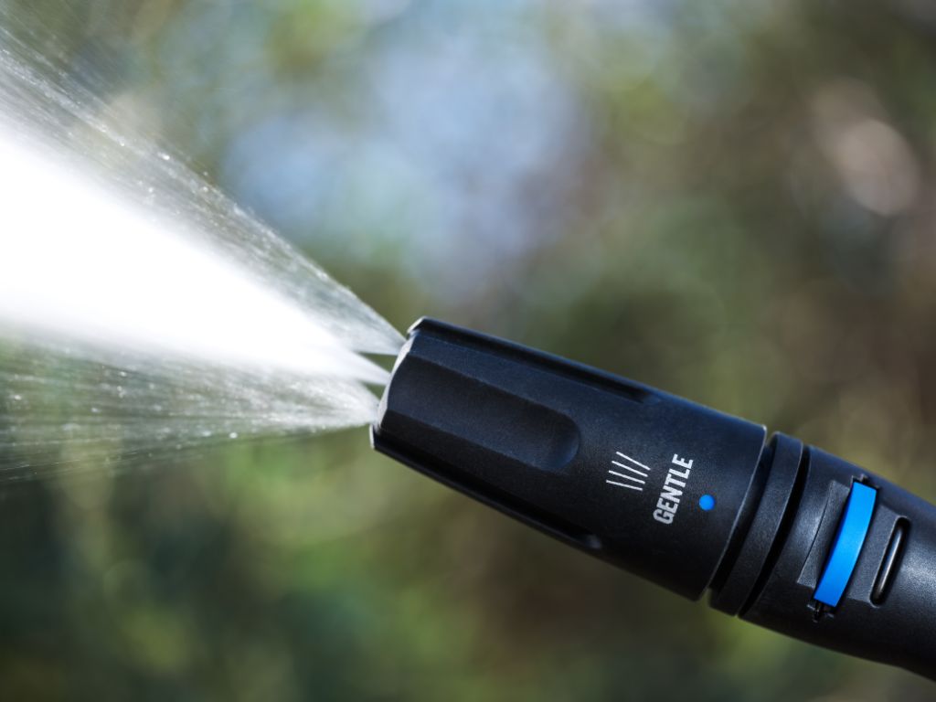 Black high-pressure cleaner by ES Core, spraying water from a nozzle with a green and brown blurred background.