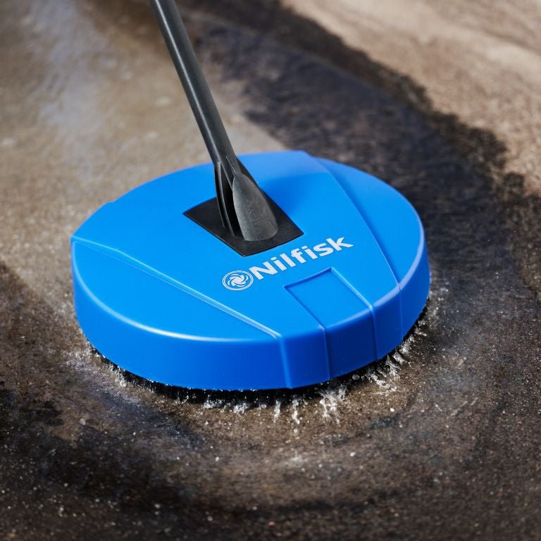Blue pressure washer brush by Nilfisk cleaning dirty concrete surface with a dark handle.

