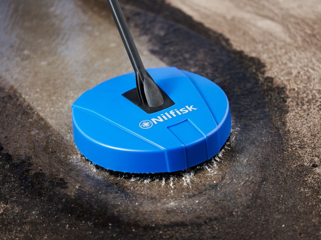Blue pressure washer brush by Nilfisk cleaning dirty concrete surface with a dark handle.
