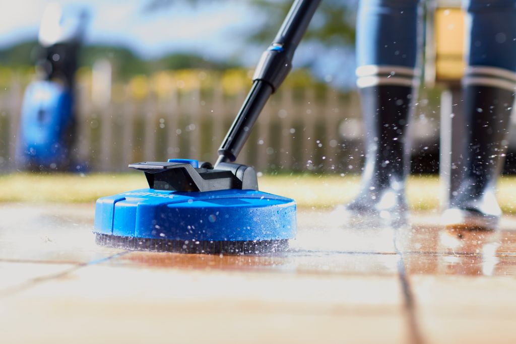 Blue patio cleaner by Nilfisk with a black handle cleaning a tiled surface, with a person in dark rubber boots standing nearby.