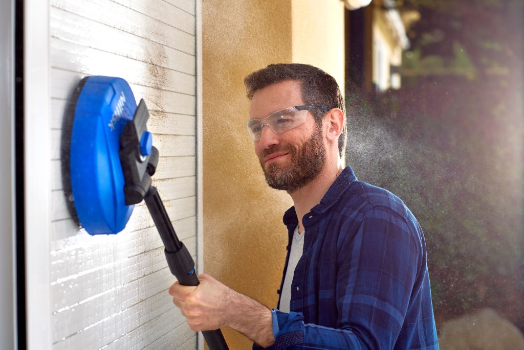 Blue patio cleaner by UK Mid used on an exterior wall with a bearded man in safety glasses.