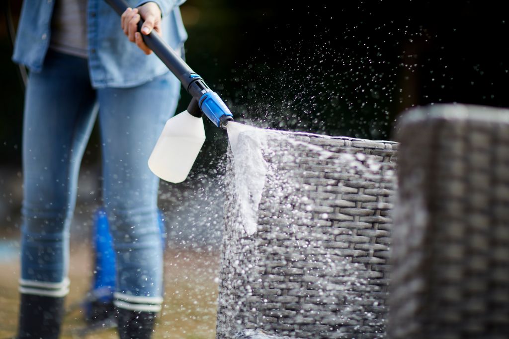 High-pressure washer cleaning kit with foam attachment by, cleaning woven outdoor furniture and being used by a person in jeans.
