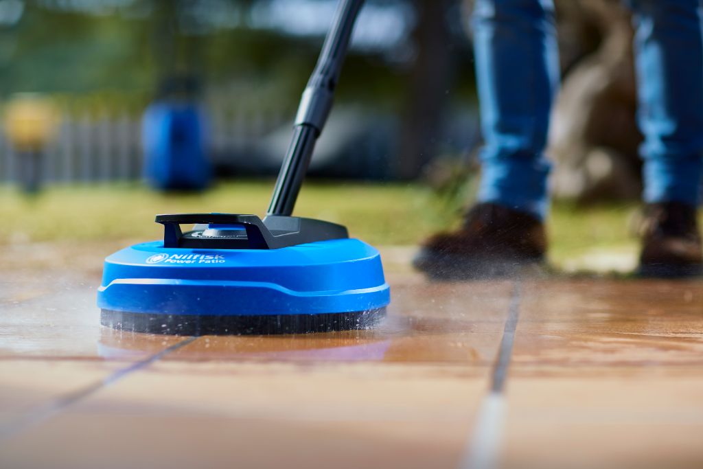Blue patio cleaner attachment for pressure washers by Nilfisk, cleaning a tiled patio surface with water jets.
