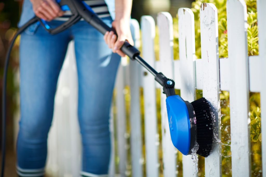 High-pressure washer attachment with rotating brush by ES, washing a white picket fence.