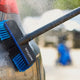 Blue and black car wash brush by Nilfisk with water spraying on a gray car.