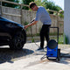 Blue pressure washer by Nilfisk being used to wash a black car, outdoors on a paved driveway.