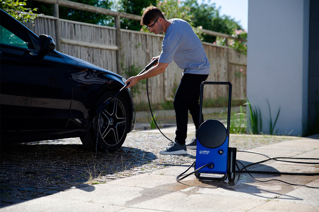 Blue pressure washer by Nilfisk being used to wash a black car, outdoors on a paved driveway.