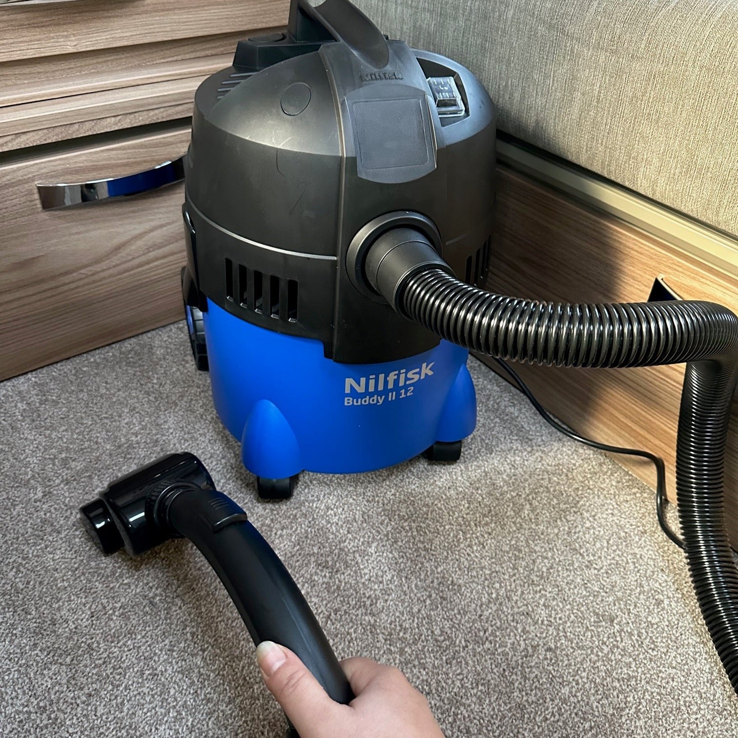 Blue wet/dry vacuum by Nilfisk with a black hose and cleaning head on a carpeted floor.
