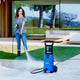 Blue pressure washer by Nilfisk being used on a light concrete patio near grass.