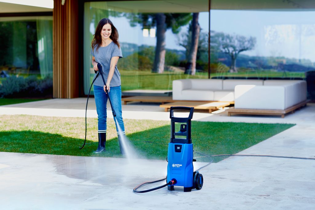 Blue pressure washer by Nilfisk being used on a light concrete patio near grass.
