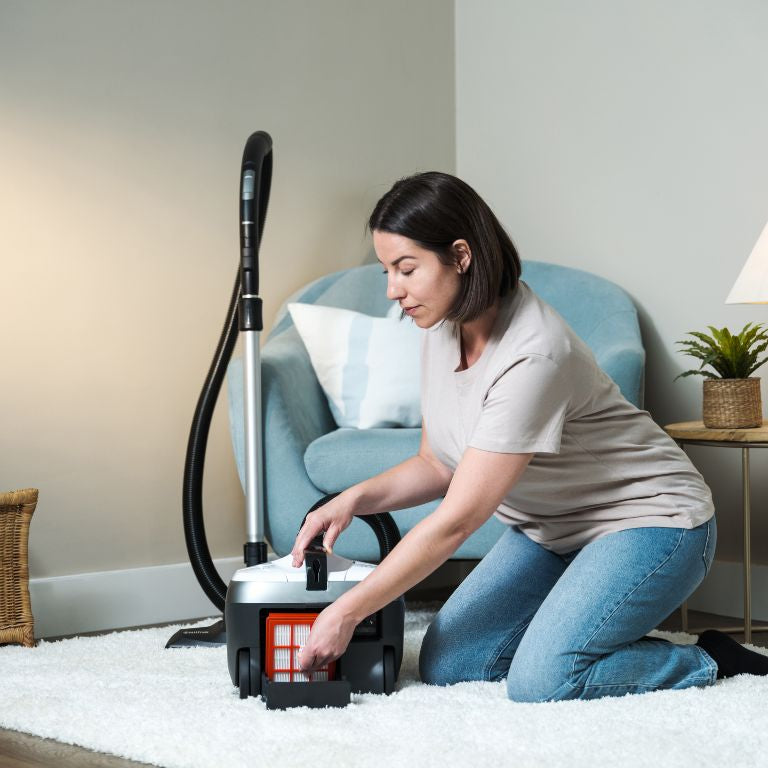 White vacuum cleaner, 650 W, beside a woman on a white rug in a living room setting.