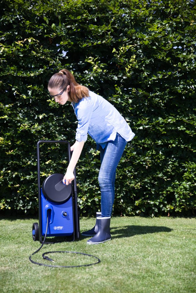 Blue pressure washer by Nilfisk with a hose reel, beside a woman in jeans and boots on grass.