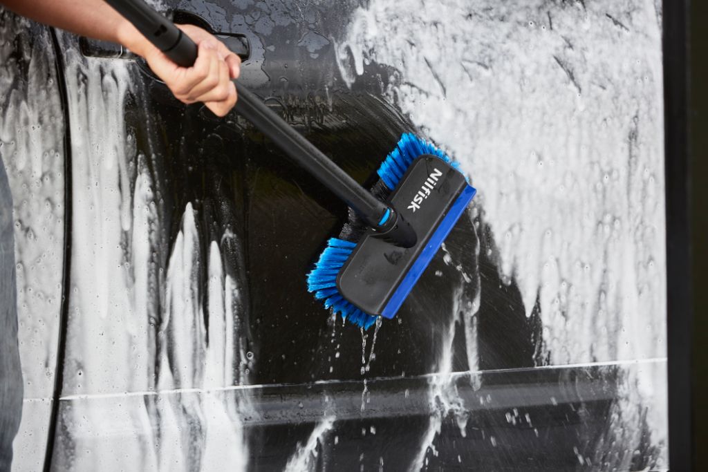 Car cleaning brush with blue bristles by Nilfisk scrubbing a soapy black car door.
